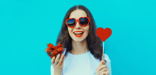 Poster - Portrait of happy young woman with strawberries holding sweet lollipop in red heart shaped glasses