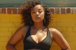 © ArtMajestic - Confident black African American curly haired woman with stretch marks under her arms, in black sports bra poses against yellow brick wall. Concept of body positive, self confidence and empowerment