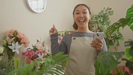 Poster - Smiling young hispanic florist confidently points to the side with her finger while holding a smartphone at her joy-filled flower shop