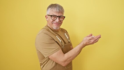 Wall Mural - Excited middle-aged, grey-haired man in glasses and apron happily pointing aside, standing with open palms, presenting an ad. astounding ad space on isolated yellow background!