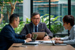 © ty - Middle aged asian businessman leading a meeting with two colleagues in a modern office in a discussion with laptop and notes on the table