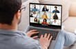 © Prostock-studio - A young man wearing glasses sits on a sofa and takes part in a virtual team meeting via a laptop with eight colleagues appearing on the screen, during the afternoon.