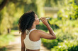 © Prostock-studio - A young woman stays hydrated by drinking water from a bottle while exercising in a green, sunlit park on a beautiful morning.