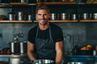 © forenna - A professional photoshoot for a food magazine featuring a handsome man in a black apron, cooking in a sleek, minimalistic kitchen. He is smiling at the camera, with large stainless