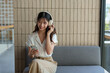 © Phimwilai - Happy Asian woman listening to music on mobile phone while sitting on sofa at coffee shop Relaxed smiling young woman with headphones in the morning Holiday relaxation concept.