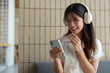 © Phimwilai - Happy Asian woman listening to music on mobile phone while sitting on sofa at coffee shop Relaxed smiling young woman with headphones in the morning Holiday relaxation concept.