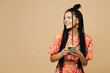© ViDi Studio - Young smiling happy Latin woman she wear orange casual clothes hold in hand use mobile cell phone look aside on area isolated on plain pastel light beige background studio portrait. Lifestyle concept