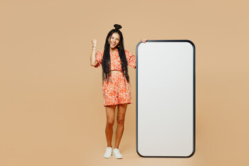 Wall Mural - Full body young Latin woman she wears orange dress casual clothes big huge blank screen mobile cell phone smartphone with area do winner gesture isolated on plain beige background. Lifestyle concept.