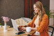© ViDi Studio - Young cheerful smiling happy IT woman wears orange shirt hold use laptop pc computer sit alone at table in coffee shop cafe restaurant indoors work or study. Freelance mobile office business concept.