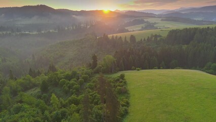 Fototapete - Aerial shot of sunrise in green mountains. Sun comes out from behind the mountain, illuminating green forest and meadows. Beautiful nature landscape