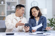 © Liubomir - Smiling couple managing their finances at home, discussing budget plans with a smartphone and paperwork. Concept of teamwork, financial planning, and domestic lifestyle.