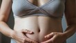 © Tom - Close-up of a pregnant woman's belly, cradled by her hands, wearing a comfortable gray top, emphasizing warmth and motherhood.