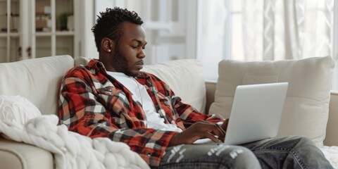 Wall Mural - Man with short curly hair wearing a red plaid shirt, using a laptop while sitting on a couch in a cozy living room.