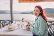 © EdNurg - Against the backdrop of the sea or lake, a woman indulges in a luxurious seafood dish, relishing the flavors of bouillabaisse on a sunny terrace.