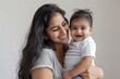 © SHI - A blissfully smiling Indian mother cradles a baby boy in a white T-shirt, grey top and jeans at home. White background, white walls