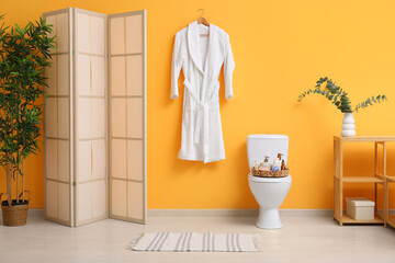 Wall Mural - White bathrobe hanging on yellow wall and different bath supplies on toilet bowl in bathroom