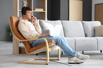 Poster - Young man with headset and laptop talking in armchair at home