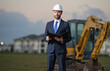 © Olena - Construction business owner. Man in suit and hardhat halmet at building construction site. Civil engineer worker in front of house near excavator. Architect, supervisor investor, construction manager.