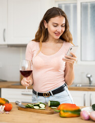 Wall Mural - Cheerful woman chatting with friends on smartphone and drinking wine while standing near table in cozy home kitchen at weekend