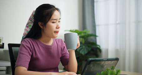 Portrait of Young asian businesswoman sitting at desk drink coffee while having a video conference on digital tablet at home office, online meeting for remote work