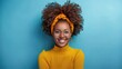 © Ai-Pixel - Portrait of a cheerful woman with curly hair and a headband, smiling against a blue background and wearing a yellow sweater.