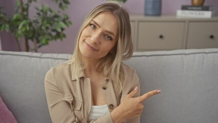 Canvas Print - Young blonde woman sitting on sofa pointing with hand finger to the side showing advertisement, serious and calm face at home