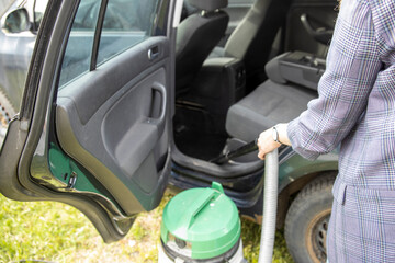 A woman is vacuuming the interior of her car. The car door is open and a green vacuum cleaner is visible. The woman is wearing a plaid shirt.