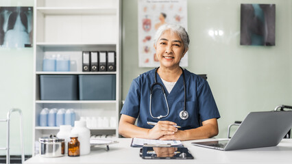 Wall Mural - A mature female healthcare professional, confidently seated at her office desk in a hospital clinic, smiles as she works on her laptop, providing expert care to patients.