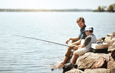 Wall Mural - Happy father, son and learning with fishing rod on rock by lake, ocean or beach together in nature. Dad with young child, kid or little boy for bonding, teaching tips or lesson to catch by sea water