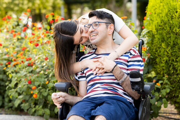 An adult man with a disability sits in an automatic wheelchair with a young woman in a park at sunset.The woman kisses the disabled man on the face.Unconditional support for people with disabilities.