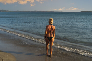 Wall Mural - A slender blonde girl in a bikini is alone on a sandy beach in Athens, Greece on a summer day, photo at sunset from the back.