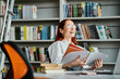 © LIGHTFIELD STUDIOS - A redhead female tutor teaches online, seated at a library desk engrossed in reading a book after her after school lesson.