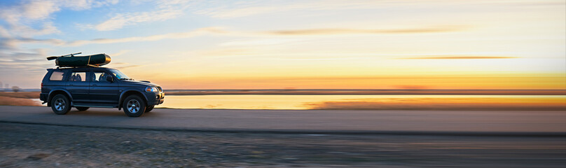 SUV with a kayak on the roof rack driving along a scenic lakeside road at sunset, reflecting an adventurous and outdoorsy lifestyle.