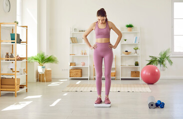 Wall Mural - Woman keeps fit with regular sports workouts. Beautiful slim young girl in pink sportswear looks down at her weight as she stands with hands on hips on scales in living room with fitness equipment