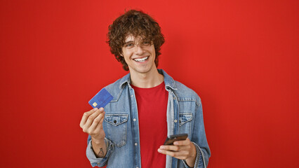 Handsome young man with curly hair smiling, showing credit card and holding smartphone, against a red background.