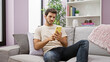 © Krakenimages.com - A handsome young man with a beard using a smartphone while sitting on a gray couch in a modern living room.