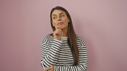 Wall Mural - A contemplative young hispanic woman in a striped shirt poses against a soft pink background, embodying beauty and elegance.