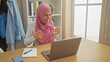 © Krakenimages.com - A cheerful woman in a pink hijab laughing during a video call on a laptop at her wooden desk inside a bright room.