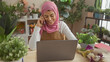 © Krakenimages.com - Smiling woman wearing hijab talking on phone while using laptop indoors surrounded by plants.