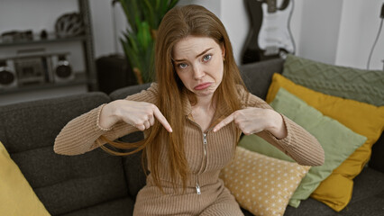 Sticker - A young blonde woman in a beige outfit looks annoyed, pointing at herself while sitting on a gray couch indoors