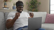 © Krakenimages.com - Happy young african american man with a beard celebrating in his living room, sitting on a sofa with a laptop on his lap, amidst cozy home decor and indoor plants.