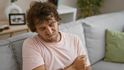 Sticker - A young hispanic man showing pain indoor, sitting in a living room with a distressed expression on his face.