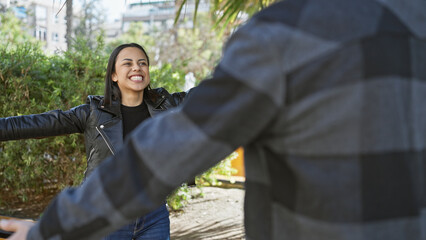 Wall Mural - A smiling woman in a leather jacket extends her arms to a blurred man on a sunny urban street.