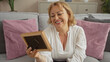 © Krakenimages.com - An elderly caucasian woman with blonde hair and a beautiful smile, sitting in her living room holding a photo frame, surrounded by pink and gray cushions on a cozy couch.