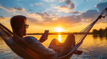 Sticker - Man relaxing in a hammock by the lake, using a smartphone during a serene summer sunset.