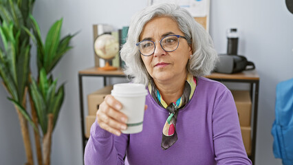 Canvas Print - Mature woman with grey hair drinking coffee in a modern office, wearing glasses and a purple sweater, exuding professionalism and confidence.