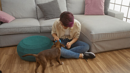 Poster - A young caucasian woman sits on the floor in her living room playing with her dog, creating a warm and cozy indoor scene.