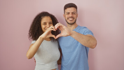 A smiling man and woman make a heart shape with their hands against a pink background, depicting affection and unity.