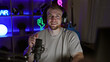 © Krakenimages.com - A handsome man with a beard, wearing headphones, smiles while hosting a podcast in a neon-lit gaming room at night.
