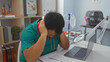 © Krakenimages.com - A stressed young chinese man in a clinic wearing a green uniform, sitting with his head in hands next to a laptop and medical paperwork.